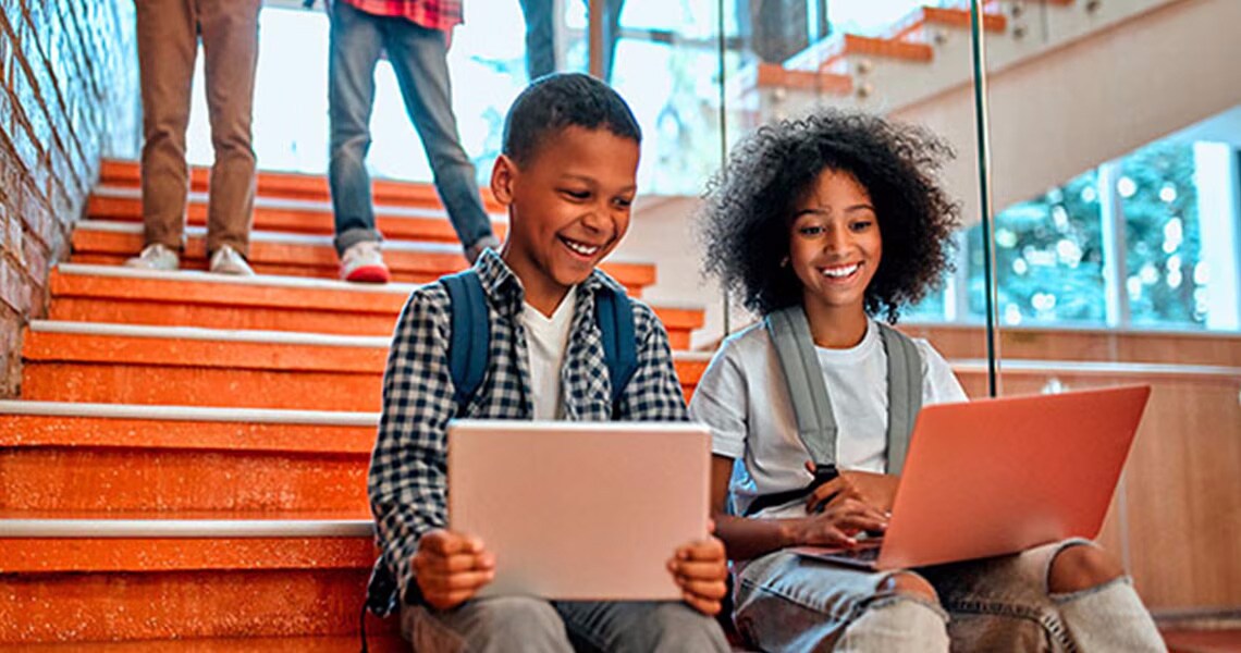 Two children sitting on the stairs and smiling while working on a laptop