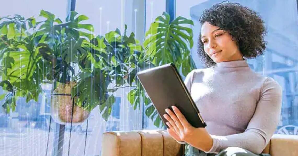 A woman sitting on a couch near plants and looking at a tablet, with OpenBlue graphics in the background