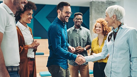 A man and a woman shaking hands, while their colleagues smile and watch around them